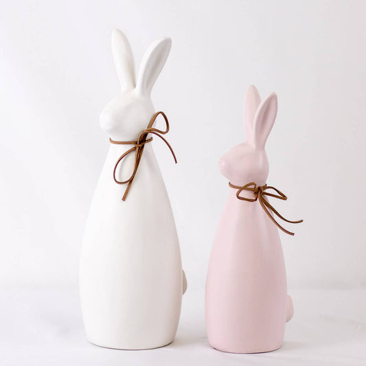 Two ceramic bunny figures, one white and one pink, tied with brown string on a light background.