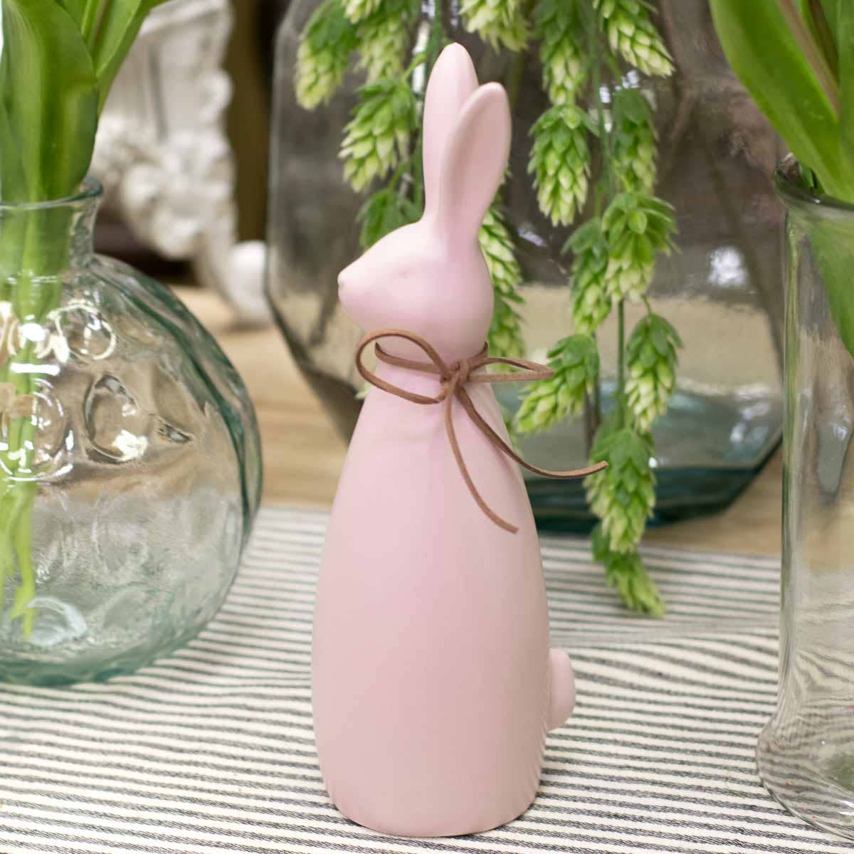 Pink rabbit-shaped bottle with a brown ribbon on a striped surface with decorative vases in the background.